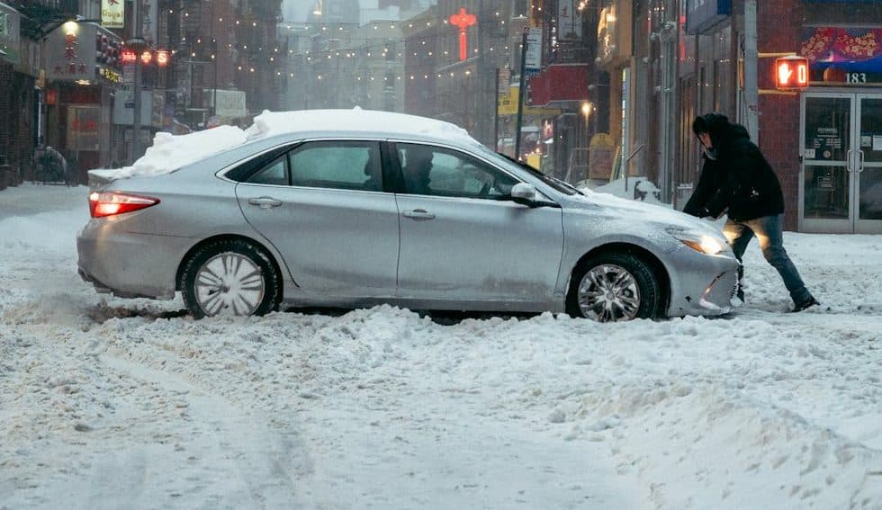Person pushing a car stuck in deep snow on a city street